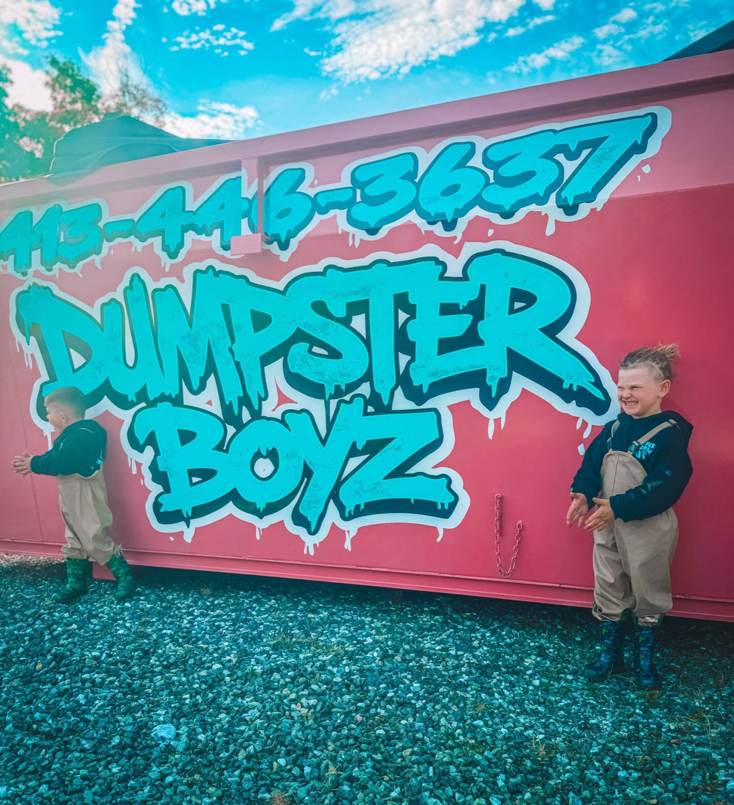 The boys laughing on either side of the giant teal Dumpster Boyz logo and phone number on a pink dumpster.